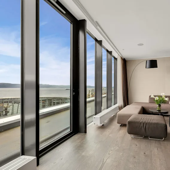 En stor hjørnesofa står i en moderne stue med gulv-til-tak-vinduer som vender mot vannet. Et lite bord med en vase blomster står foran.