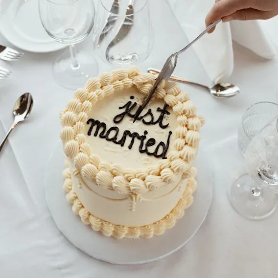 En hånd skjærer i en hvit kake med teksten 'just married', omgitt av dekket bord for en feiring.