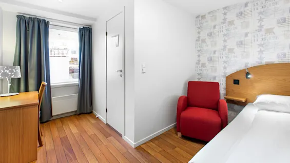 En rød lenestol står ved siden av en seng i et moderne hotellrom med tregulv. Et skrivebord, stol, lampe og et vindu med blå gardiner fullfører den minimalistiske innredningen.
