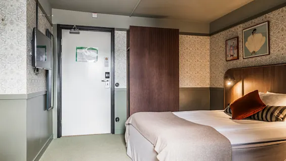 Ett mysigt hotellrum med en prydligt bäddad dubbelsäng, stämningsfull belysning, konst på väggarna och en öppen dörr som leder till korridoren.