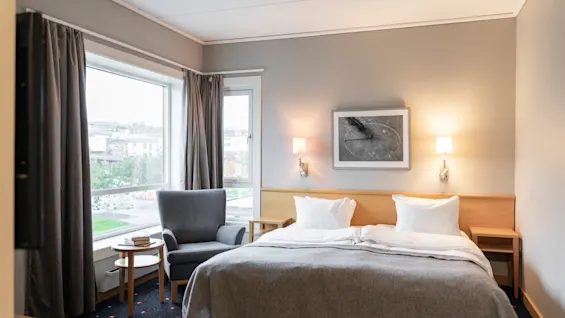 Et koselig hotellrom med en komfortabel seng, en grå lenestol ved vinduet og varm belysning, som inviterer til avslapning.