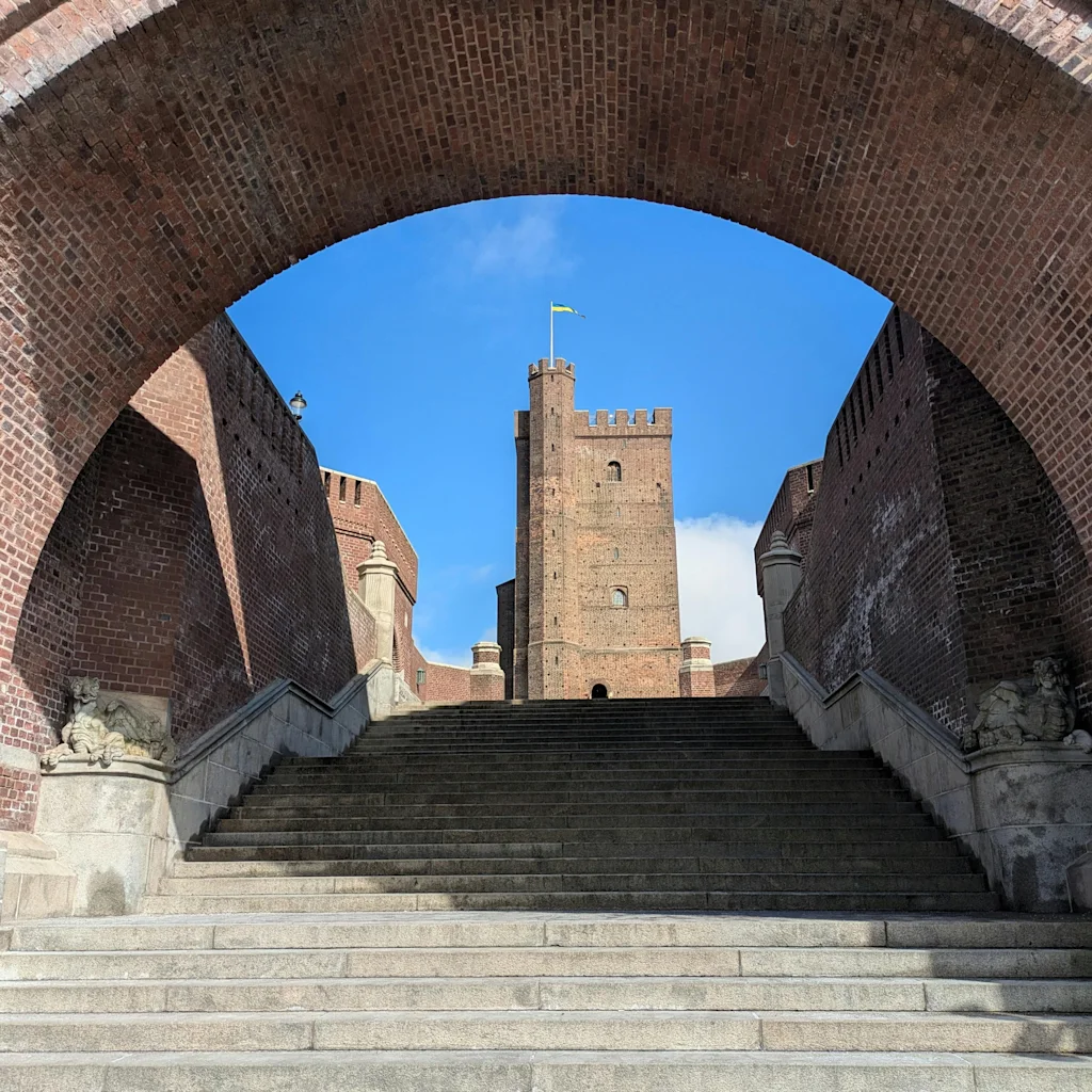 Utsikt gjennom mursteinsbue opp steintrapper til Kärnan tårnet i Helsingborg, Terrasstrapporna under blå himmel.