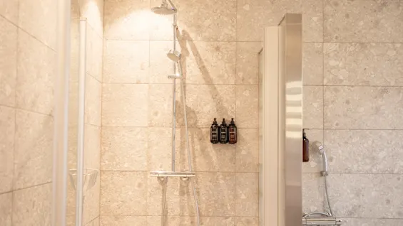 Showerhead mounted on a tiled wall, surrounded by three hanging bottles. The wall features beige, speckled tiles, creating a neutral, modern bathroom setting.