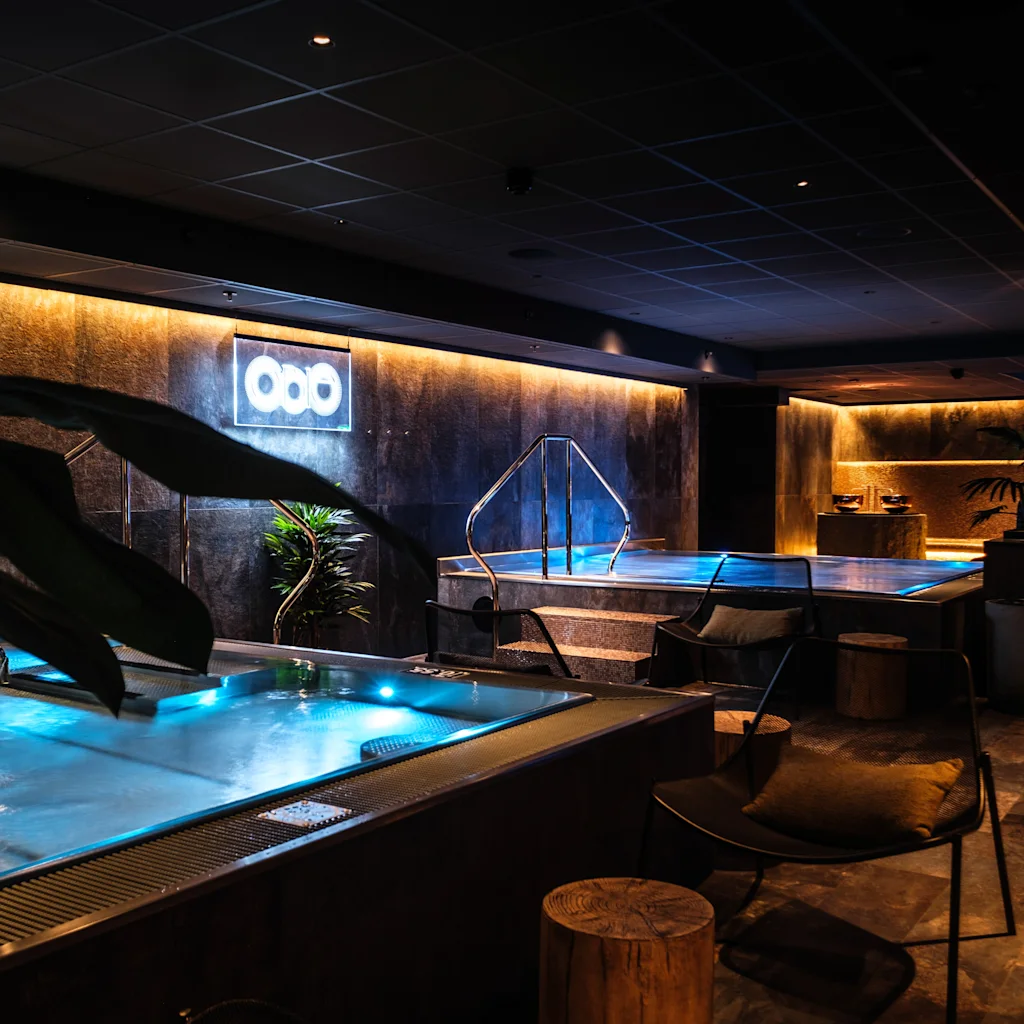 Luxurious indoor spa features illuminated pools, surrounded by ambient lighting and modern decor, including plants and seating. Neon sign with three circles on wall provides subtle illumination.