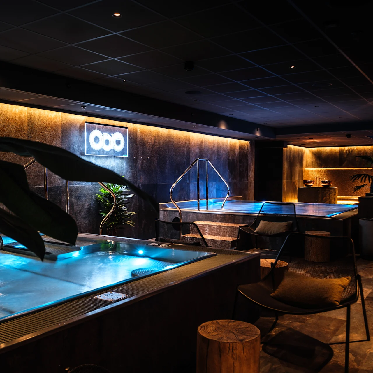Lyxigt inomhusspa med upplysta pooler, omgivna av stämningsbelysning och modern inredning, inklusive växter och sittplatser. Neonskylt med tre cirklar på väggen ger subtil belysning.