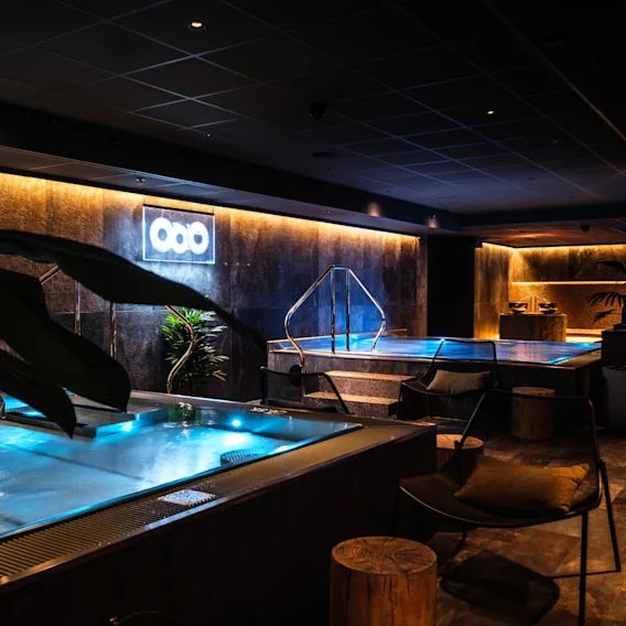 Luxurious indoor spa features illuminated pools, surrounded by ambient lighting and modern decor, including plants and seating. Neon sign with three circles on wall provides subtle illumination.