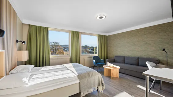 Lyst hotellrom med dobbeltseng, grå sofa, grønne gardiner og store vinduer som slipper inn dagslys.