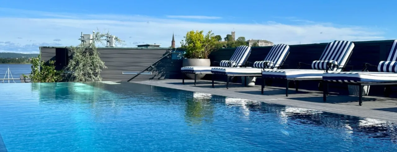 En infinitypool med solstolar på en däck, med utsikt över en stadssilhuett under en blå himmel.