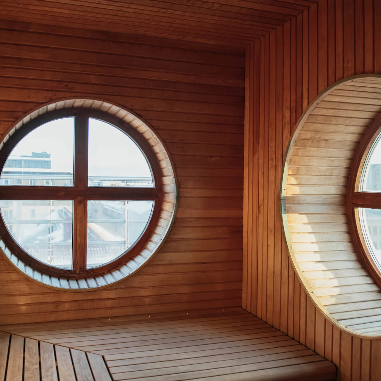 Wooden sauna interior features circular windows providing cityscape views, creating a warm, inviting atmosphere. Natural light casts shadows across the benches, enhancing the tranquil setting.