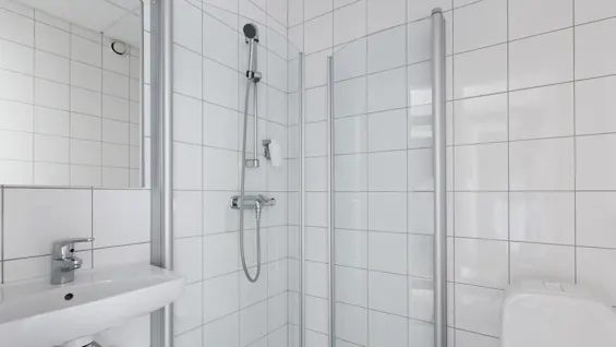 En dusjkabin med klare glassdører og et kromdusjhode er i et hvitt flislagt bad. En hvit servant med kromkran er ved siden av.