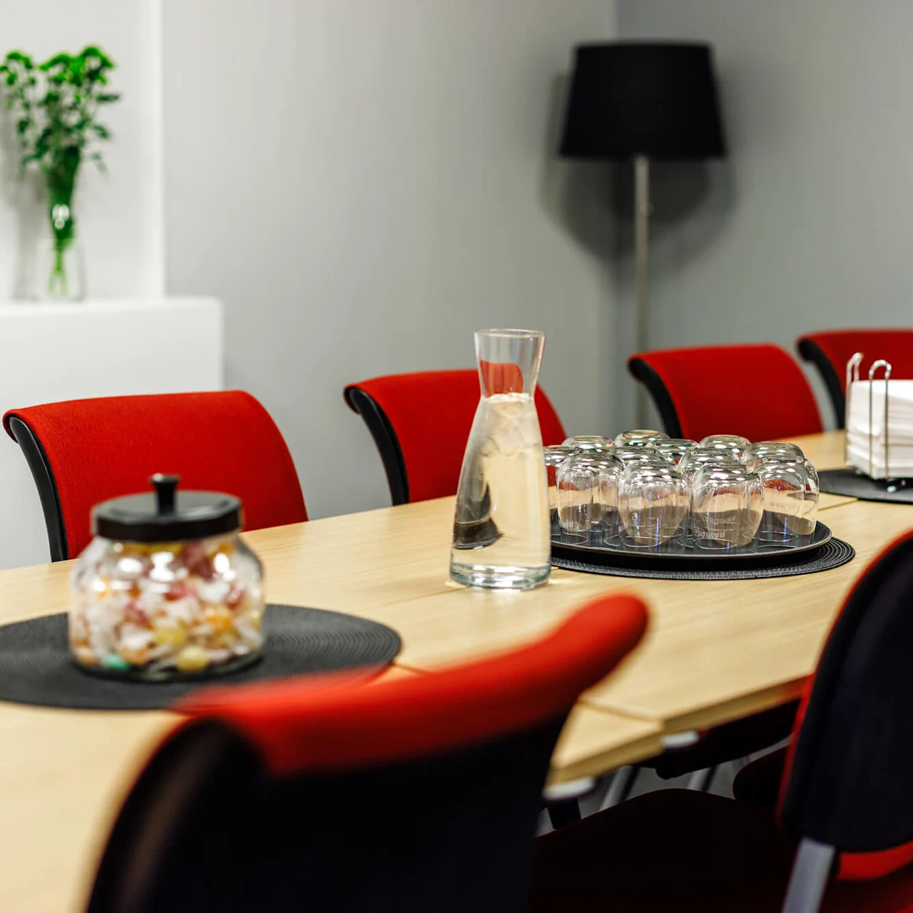 Ett långt konferensbord har en burk godis, en vattenkaraff, uppochnervända glas och servetter. Röda stolar omger bordet i ett minimalistiskt mötesrum.