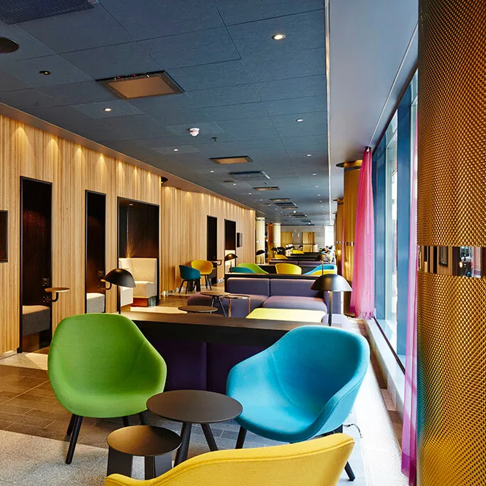 Lyse, farverige stole og sofaer står omkring små borde i en moderne lounge, med en træpanelvæg med tv'er og separate siddeområder, mens store vinduer lader naturligt lys komme ind.