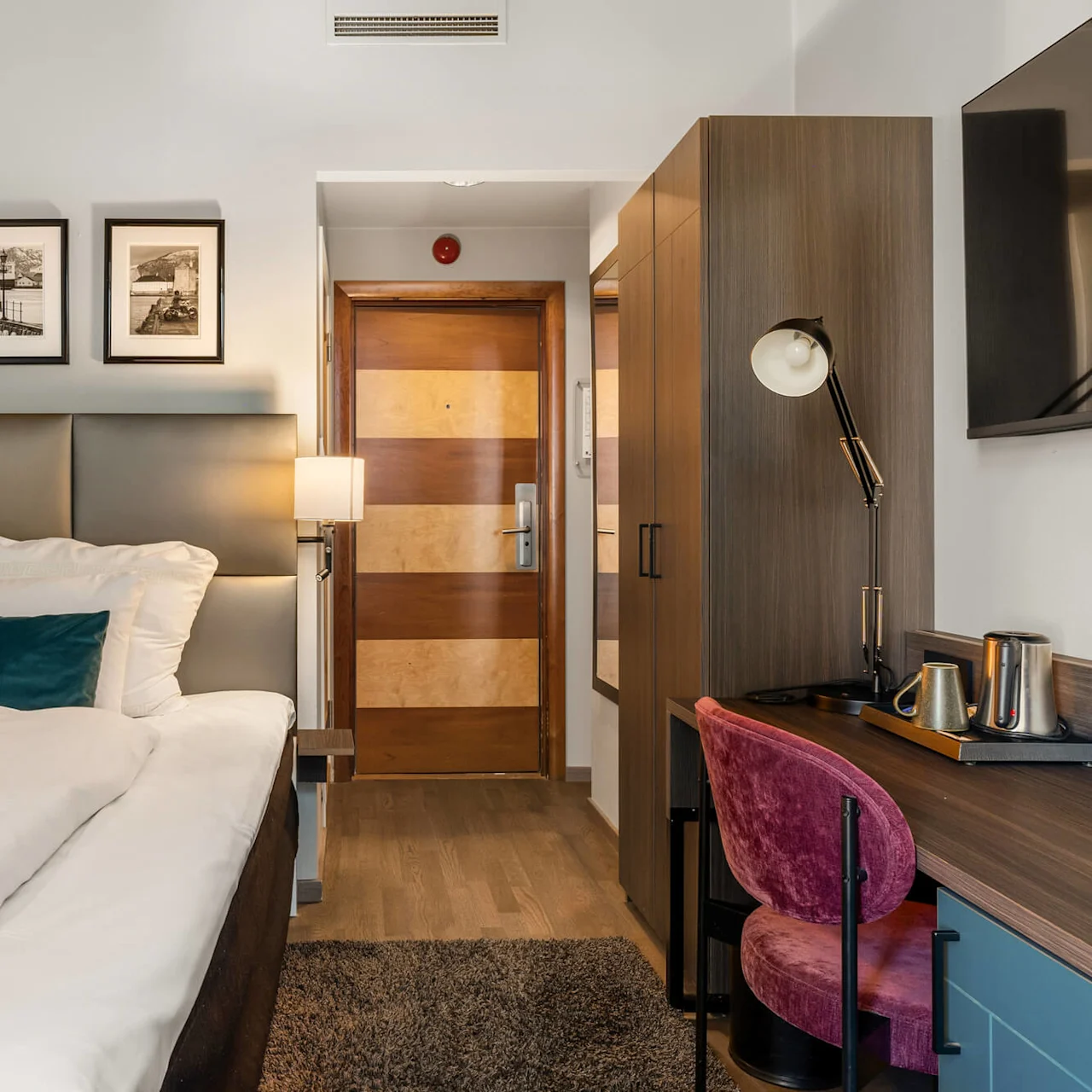 En seng med hvidt sengetøj og en turkis pude står nær en trædør på et hotelværelse. Vægkunst, et klædeskab og et skrivebord med en lampe skaber en hyggelig atmosfære.