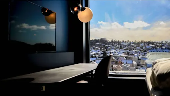 Skrivebord står ved siden af et vindue, der viser en snedækket by under en blå himmel. En moderne lampe hænger over og oplyser rummet med blødt lys.