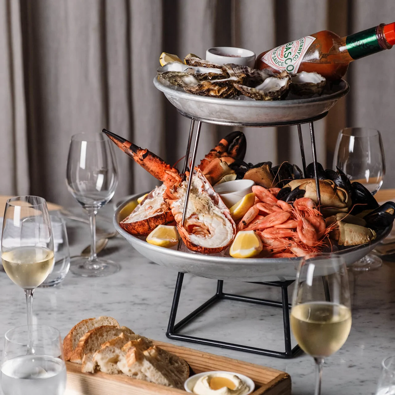 Et etasjefat med sjømat har hummer, reker, østers og sitronbåter på et marmorbord med glass hvitvin og brød. En flaske merket "Tabasco Original Red Sauce" hviler på toppen.
