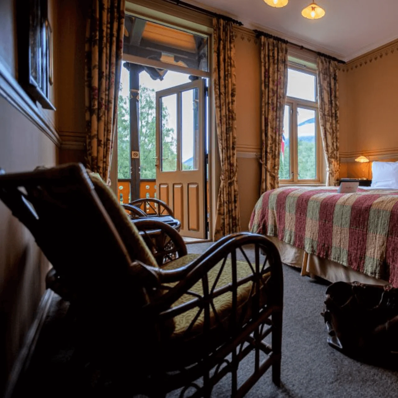 A cozy hotel room features a double bed with a patterned quilt and pillows, an open glass door leading to a balcony, and wicker chairs in a warmly lit setting.