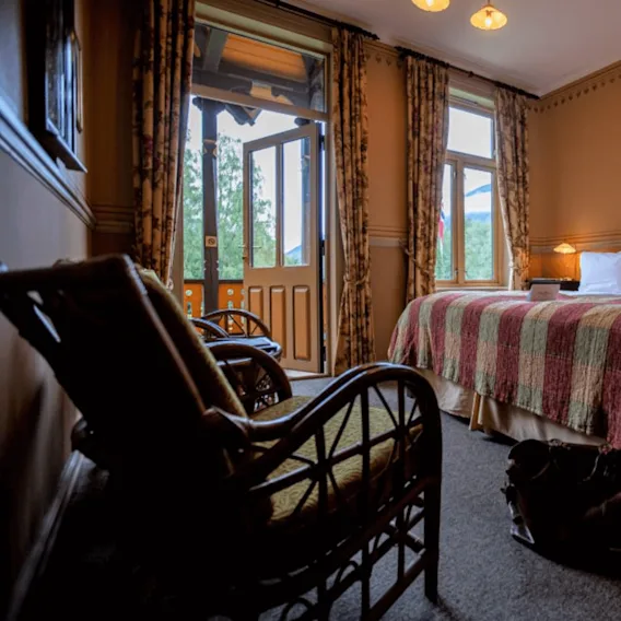Et koselig hotellrom med en dobbeltseng med et mønstret teppe og puter, en åpen glassdør som fører til en balkong, og kurvstoler i et varmt opplyst miljø.