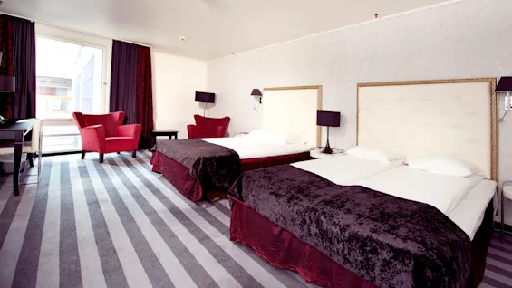 A hotel room with two beds, red chairs, and a balcony, illuminated by natural light.
