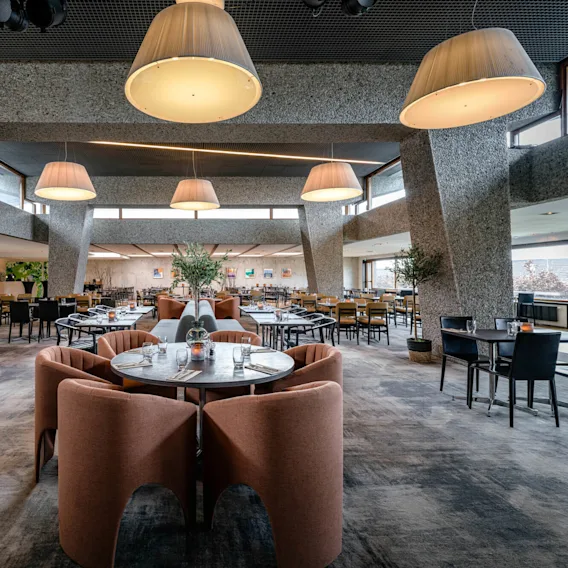 En restaurantinteriør har plysjstoler rundt runde bord. Store, moderne taklamper henger ovenfor og lyser opp det romslige området med polerte gulv og moderne dekor.