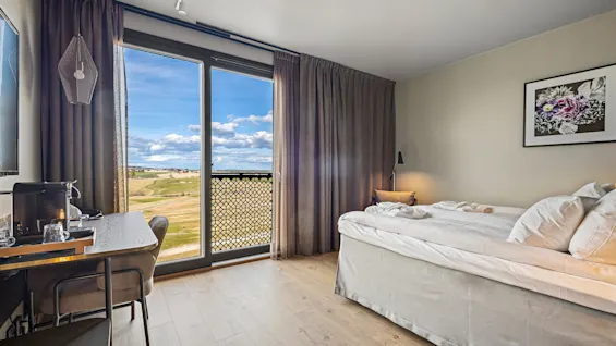 En säng står bredvid ett stort fönster med öppna gardiner, med utsikt över ett naturskönt landskap. Ett litet skrivbord med stol är placerat i närheten och skapar en bekväm rumsmiljö.