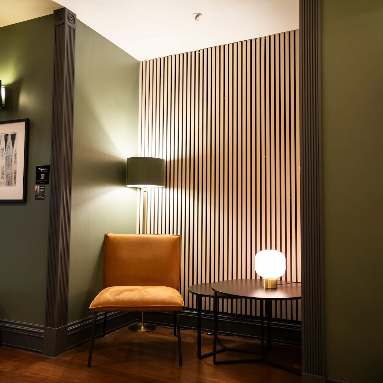 En tom sennepsgul stol står ved siden af en lampe på to borde, i et moderne rum med grønne vægge og kunstværk. Vertikale trælameller pryder en væg.