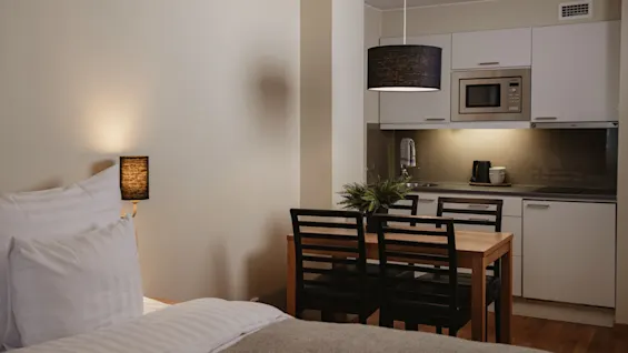 A neatly made bed faces a dining table with black chairs, situated near a modern kitchenette; soft lighting and minimal decor create a cozy atmosphere.