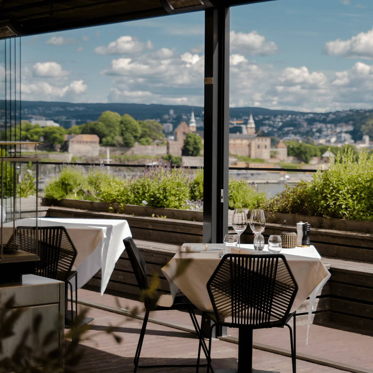 Bord dekket med vinglass og pepperkvern, plassert på en terrasse med utsikt over frodig vegetasjon og fjerne åser under en delvis skyet himmel.