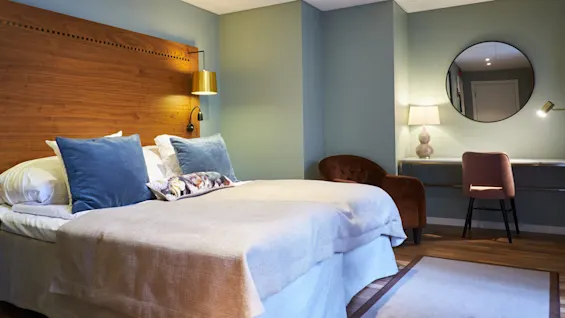 A neatly made bed with blue pillows in a cozy, teal and brown toned bedroom with wooden accents, ambient lighting, and minimalistic decor.