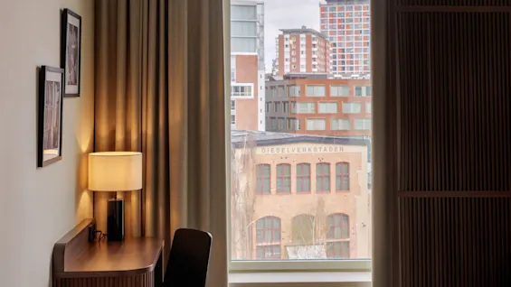 A room with a desk, lamp, and two framed pictures, viewing an urban landscape through a window; heavy curtains partially frame the scene.