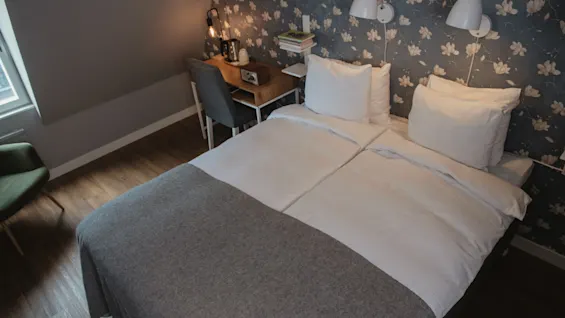 A neatly made double bed with white and gray linens, under floral wallpapered walls, is positioned next to a wooden desk with a chair, books, and a lamp.