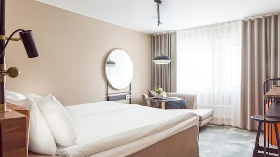 En pænt redt seng med hvide lagener står i et hotelværelse med beige vægge, et rundt spejl, et lille bord med stole og store gardiner.