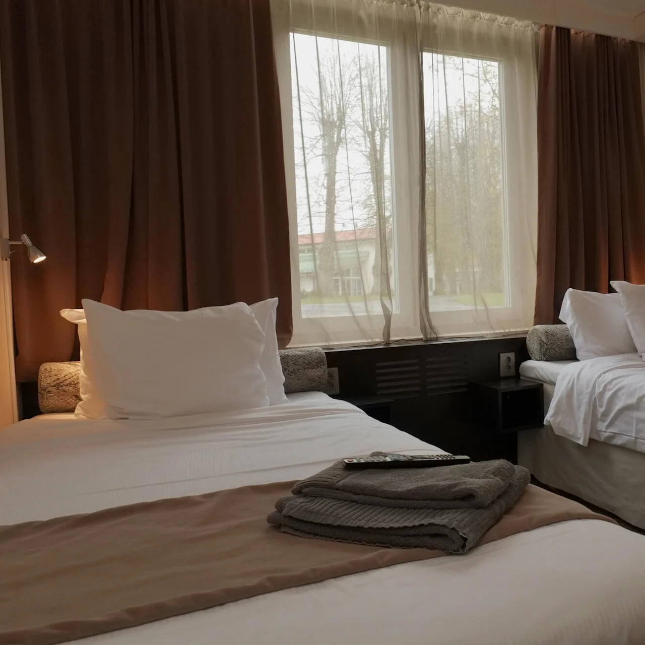 To pænt redte enkeltsenge i et hotelværelse, dækket med hvide lagner og brune tæpper, vender mod et vindue med brune gardiner. En fjernbetjening ligger på en seng.