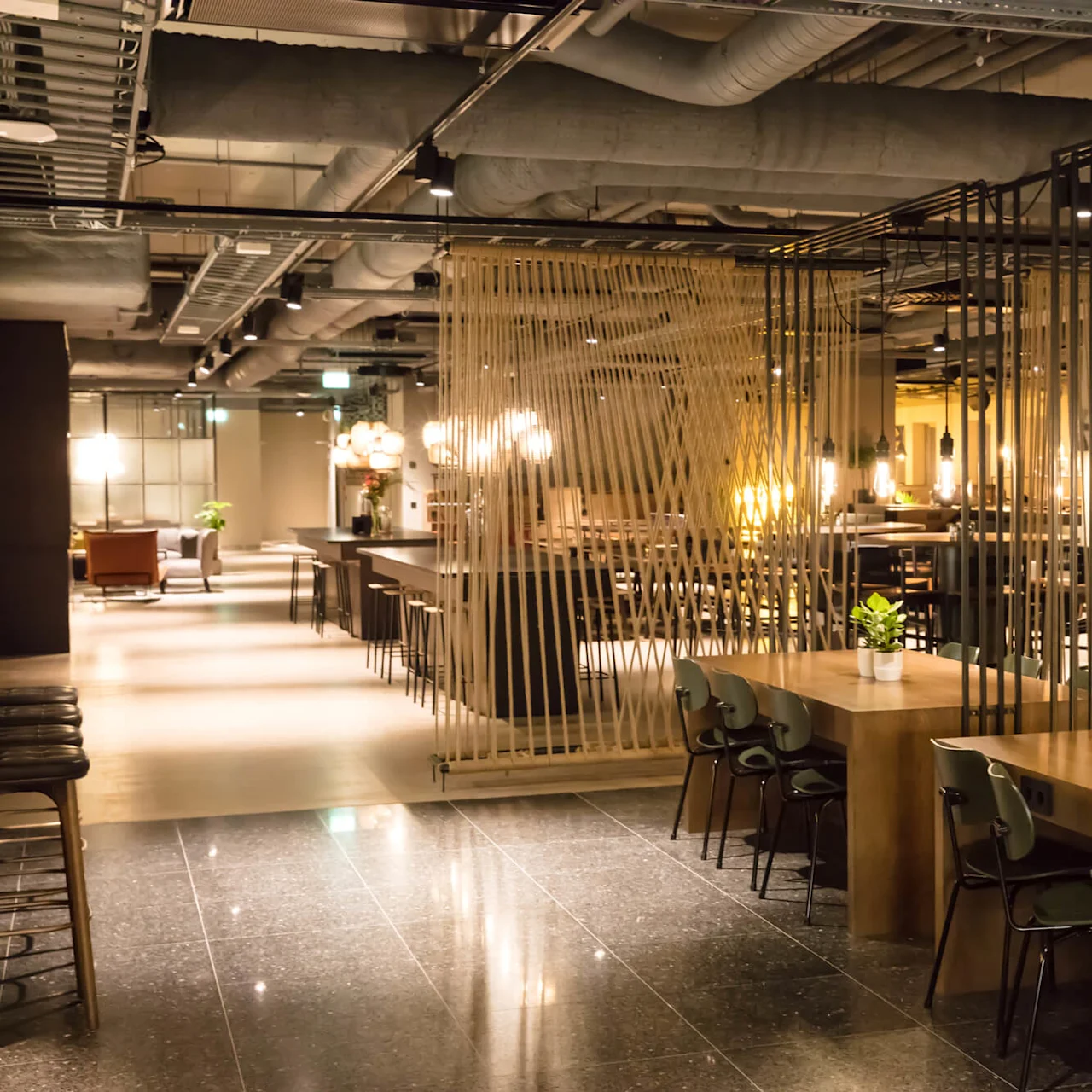 A modern restaurant with warm lighting, wood furniture, and hanging rope dividers. Exposed ceiling pipes contribute to an industrial vibe.