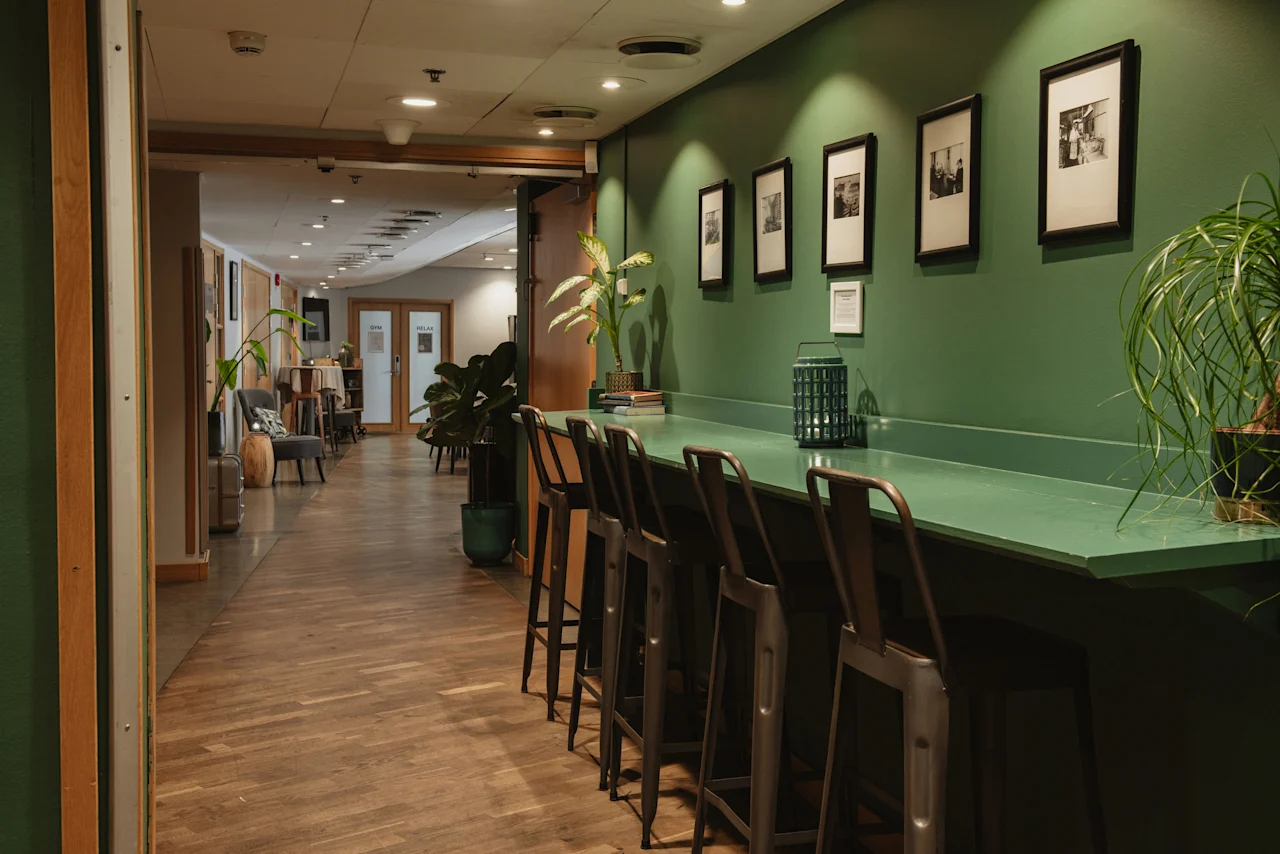 A stylish hotel hallway at Home Hotel Uppsala with a long green counter and bar stools, framed art on green walls, and lush plants.