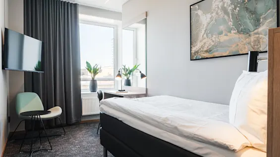 En prydligt bäddad säng med vita sängkläder står där. I närheten finns ett skrivbord med två lampor och växter vid ett stort fönster. En TV är monterad, och konstverk hänger på väggen.