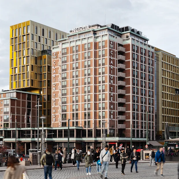 Folk går på en bygade foran en høj bygning med teksten CLARION THE HUB, omgivet af andre bystrukturer.