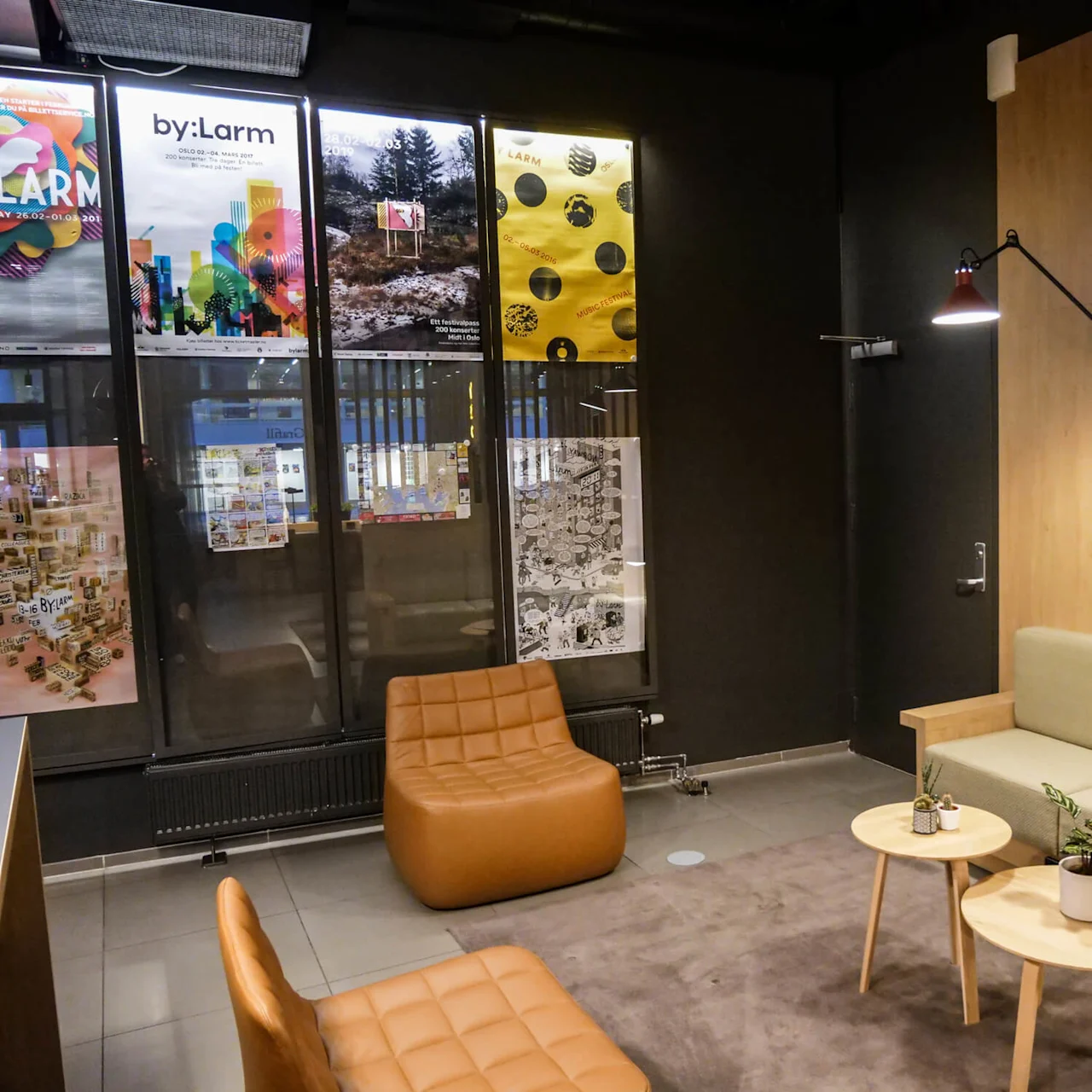 Stylish seating area features tan chairs, light wood tables with plants, sage sofa, wall-mounted lamps. Context includes posters visible through large windows. Posters read: "by:Larm," "MMXX," "2020," and dates.
