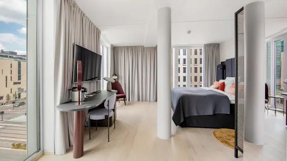 A modern hotel room with a double bed and large windows displaying an urban view. A desk with a chair and TV is located beside the bed.