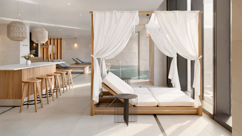 Luxurious lounge chairs rest under white drapes beside a sleek bar with wooden stools, in a modern interior bathed in natural light from tall windows.