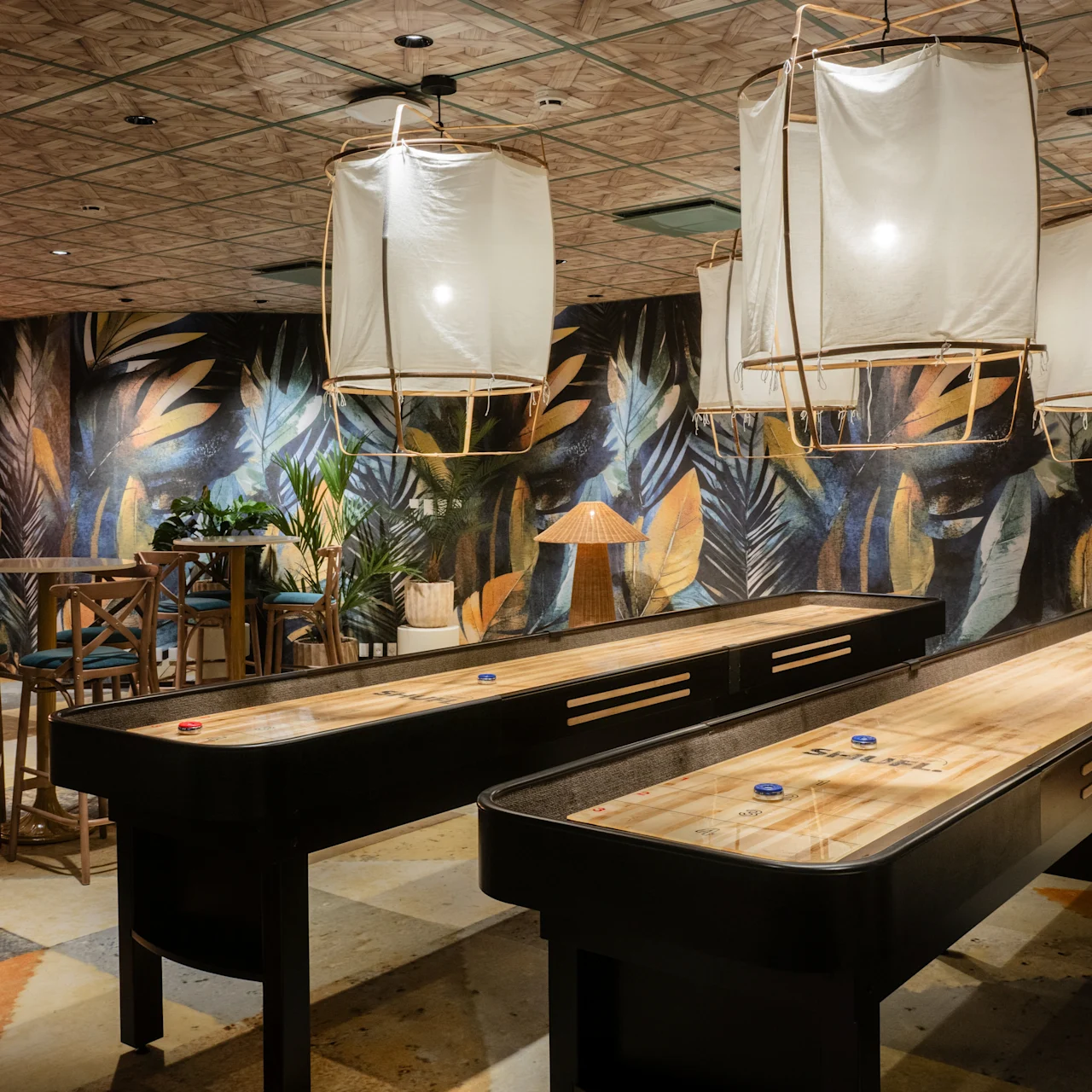 Shuffleboard tables rest in a dimly-lit room adorned with leafy murals. Overhead, large lanterns illuminate the space, which includes a dartboard and high stools around a table.