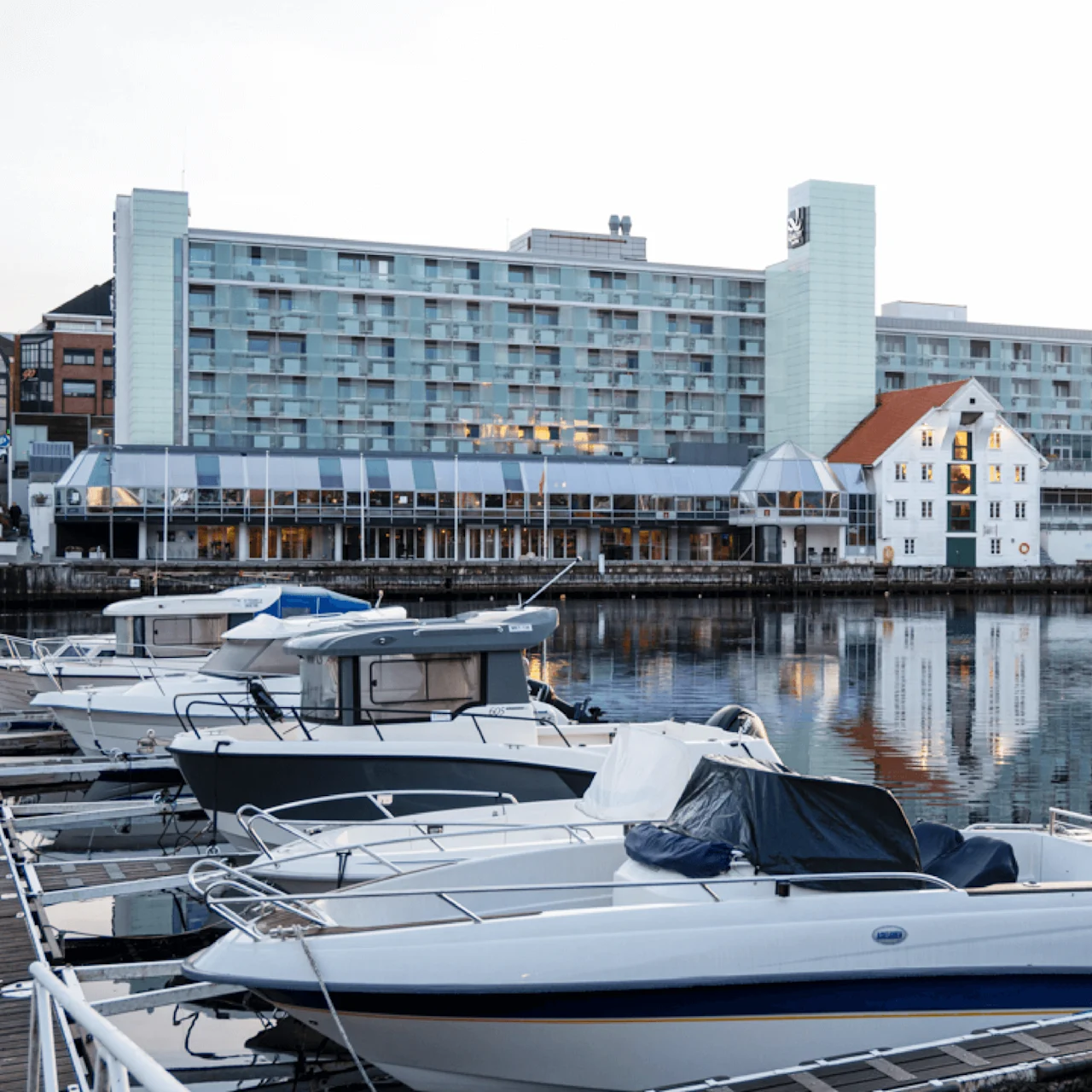 Båtar är förtöjda i en lugn marina. Ett modernt hotellkomplex står i bakgrunden. Flera flaggor syns vid hotellets entré. Vattnet speglar scenen.