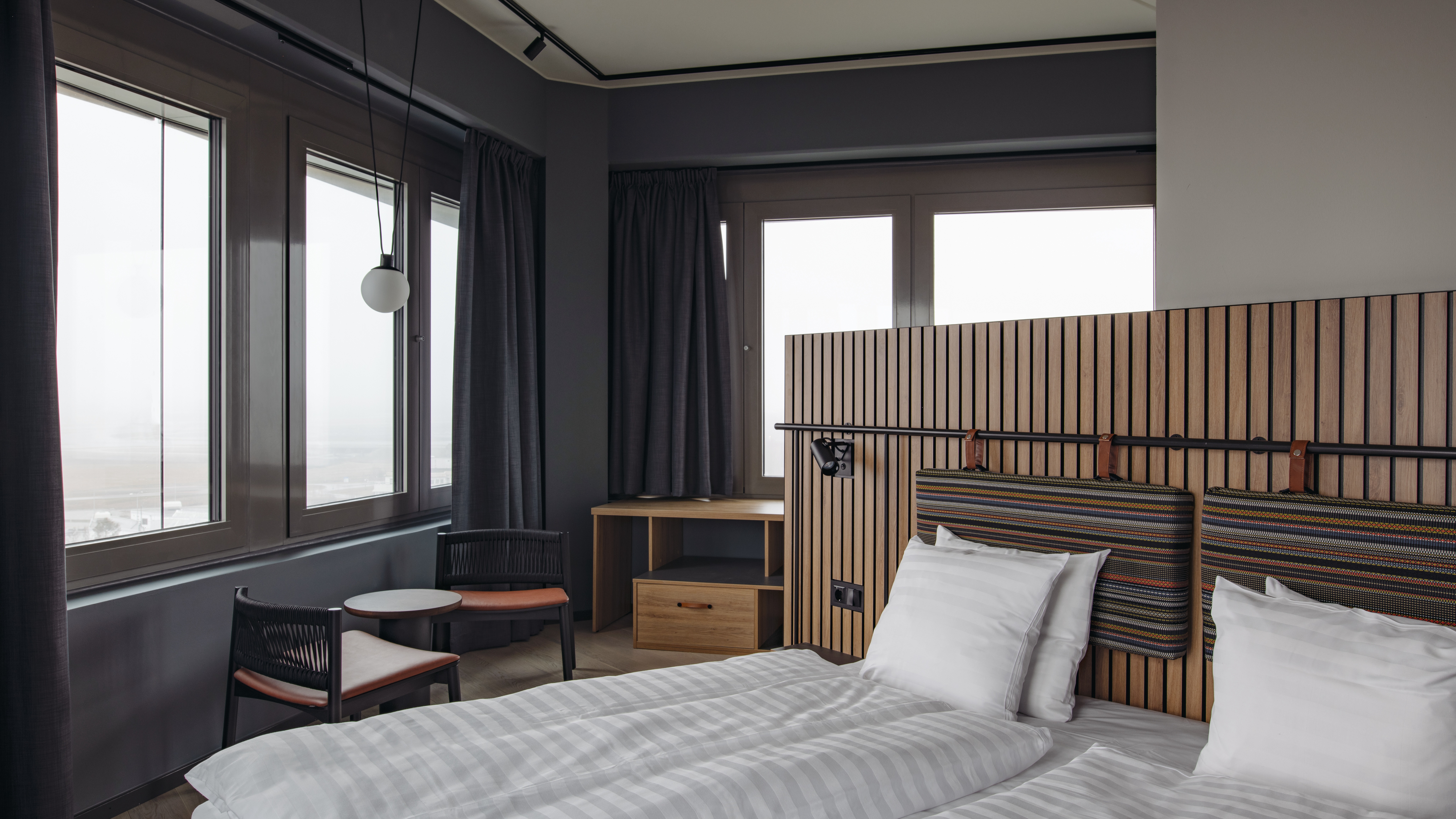 Et moderne hotelværelse med to senge, en sengegavl i træ, et lille siddeområde og store vinduer.