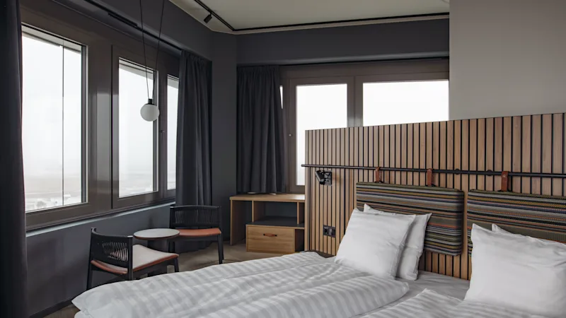 Et moderne hotelværelse med to senge, en sengegavl i træ, et lille siddeområde og store vinduer.