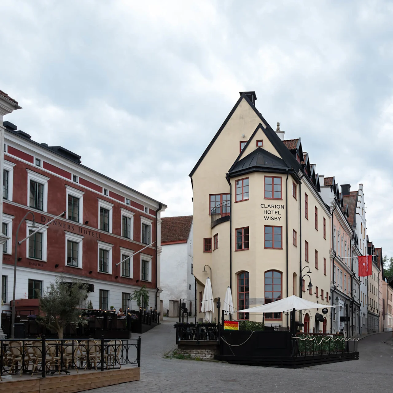 Et gadebillede viser historiske bygninger med røde og beige facader, inklusive "Clarion Hotel Wisby" og "Danner's Hotell." Udendørs siddepladser er tilgængelige under parasoller.