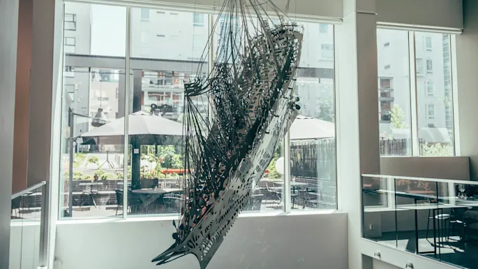 En metallisk, abstrakt skulptur henger i et moderne inneområde med store vinduer som vender ut mot en utendørspatio med bord og parasoller.