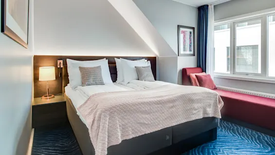 En ordentligt redt dobbeltseng med puder står under et skråt loft, ved siden af en lampe på et natbord. Et vindue med blå gardiner lyser rummet op.