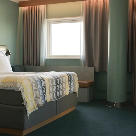 A bed, neatly made with white pillows and a patterned throw blanket, stands in a softly lit room. Nearby, curtains frame a small window, creating a cozy atmosphere.