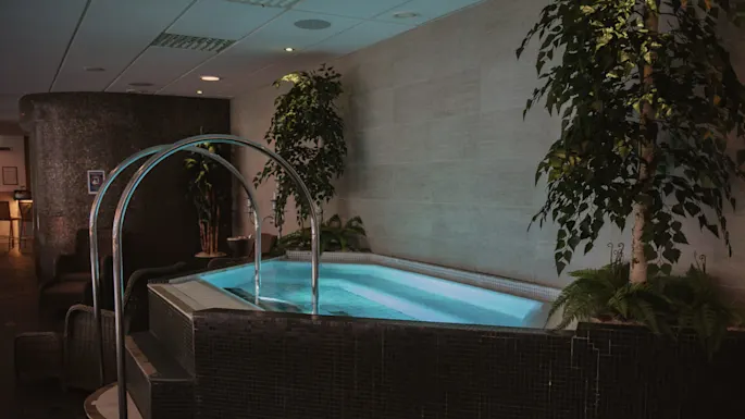 Innendørs boblebad med boblende vann og metalldetektorer, omgitt av kurvstoler og potteplanter. Myk belysning og flislagte vegger skaper en avslappende spa-atmosfære.