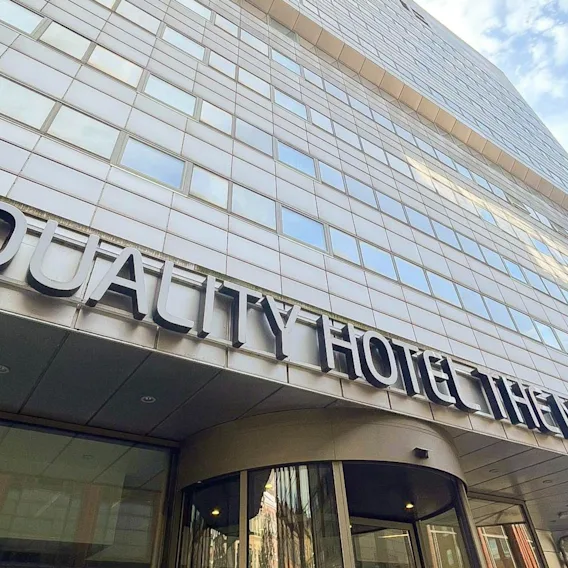 Quality Hotel The Mill sign adorns a modern building's facade, stretching above a glass entrance with revolving doors, under a blue sky.