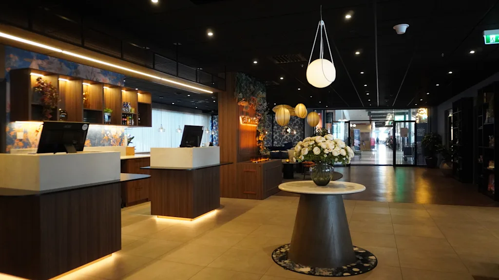 Reception desks with computers stand illuminated, surrounded by decorative hanging lights. A table with a floral arrangement is centered in a stylish lobby featuring dark wood finishes and tile flooring.